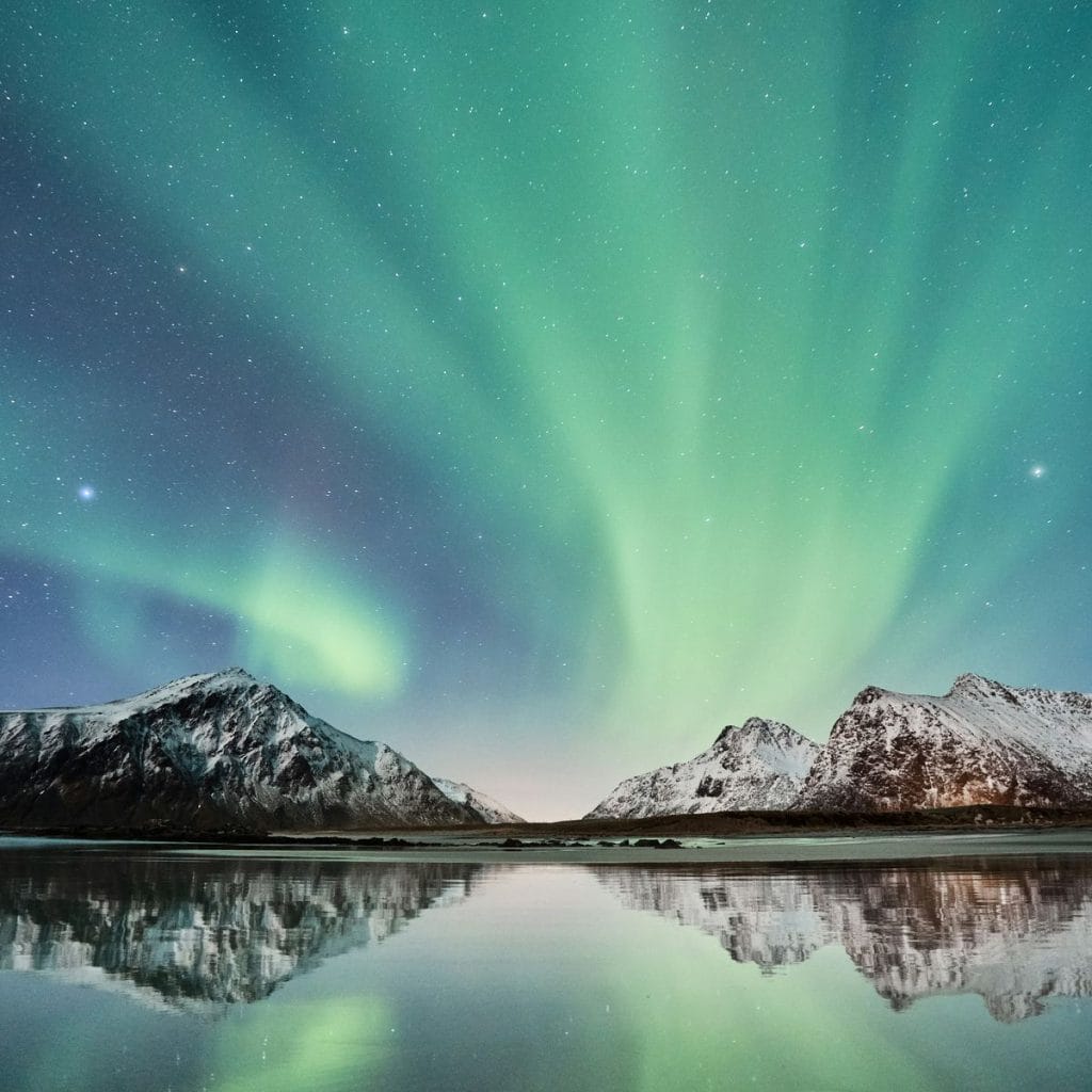 Strålende nordlys i grønt og blått over stille sjø og snøkledde fjell. Himmel med stjerner bak nordlyset.