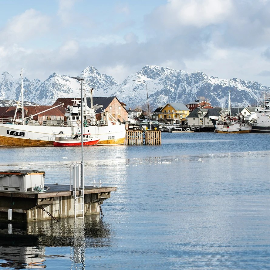 Fiskelandsby i Nord-Norge, med store og mindre fiskeskøyter. Snøkledde fjell og blå himmel bak. En stor del av norsk eksport og naturressurser.