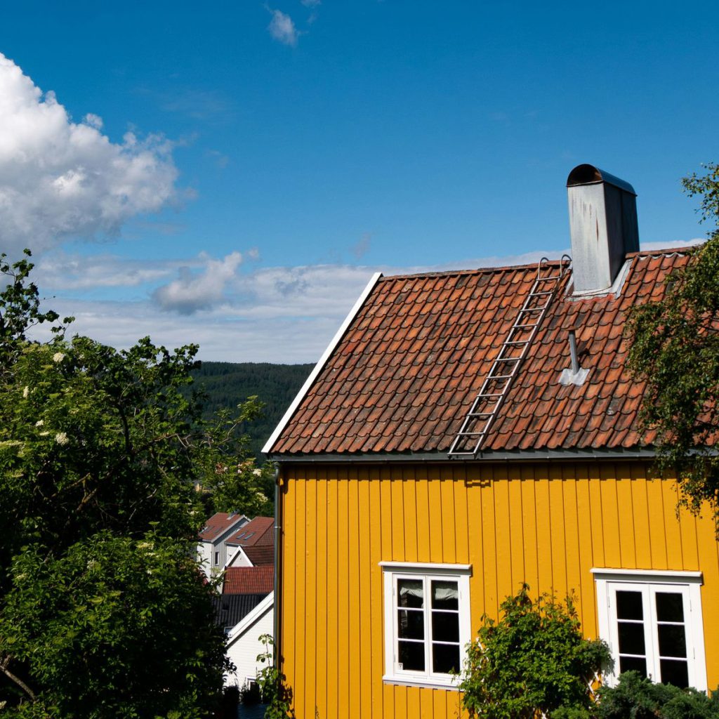 Klassisk norsk trehus i okergult, med stående panel, rød takstein og pipe. Grønne busker og blå himmel.